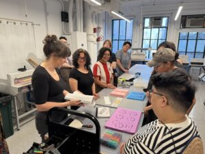 A crowd stand around a work table with printouts, dice, and craft supplies to assemble an artist's book in front of them. Elaine Lopez, the designer of the book, stands to one side explaining something as everyone listens.