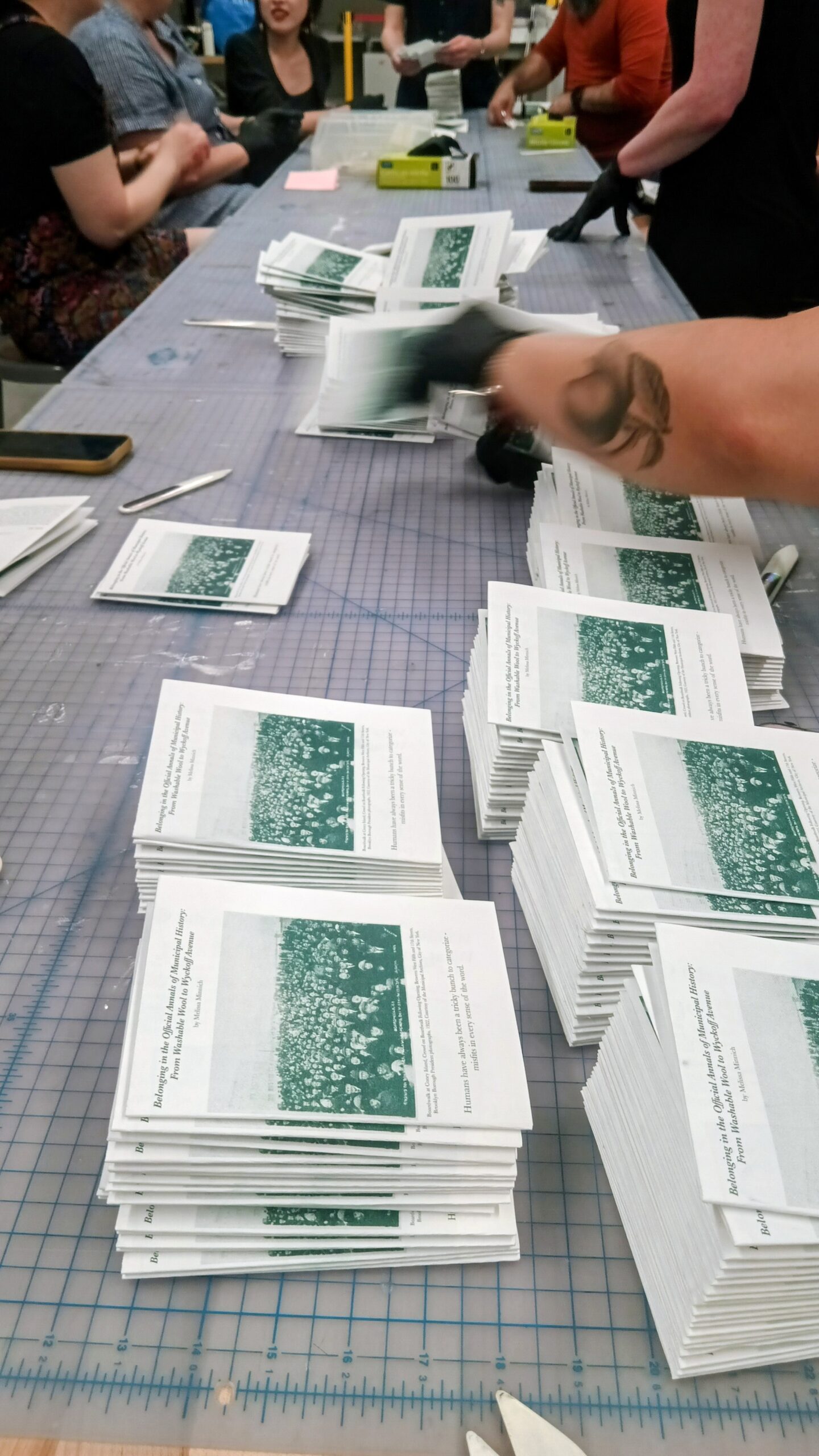 People on either side of a long table fold small white zines with vibrant green ink.