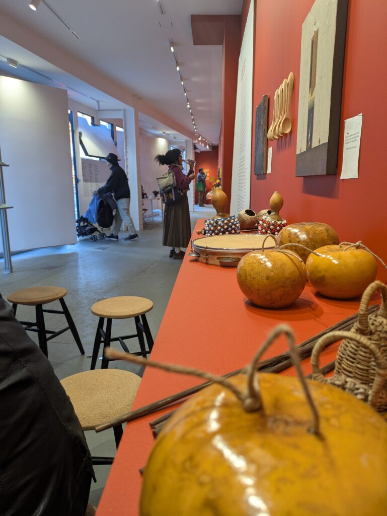 Foreground: a red shelf holding various musical instruments; wooden spoons hanging from the wall; behind: gallery visitors exploring wall displays