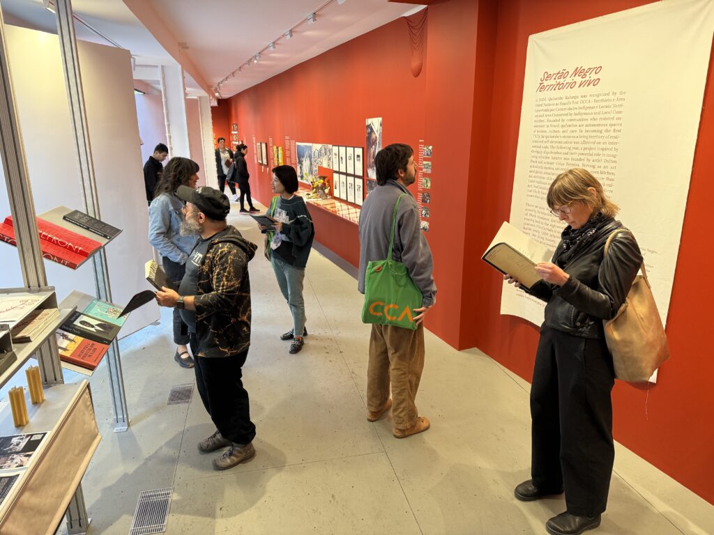 A group of people milling about an exhibition in a triagular gallery with red walls; we see shelves of books to the left and various prints and artifacts on the wall to the right