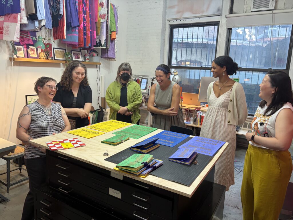 Six people stand around a tall work table with colorful paper laid out on it.