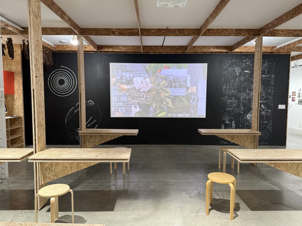 A wooden chipboard scaffolding holding several rudimentary desks, stools, a chalkboard with a complex diagram, a projected collage 