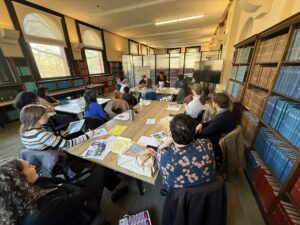 People gather around a table covered in papers with bookshelves to the right and windows to the left.