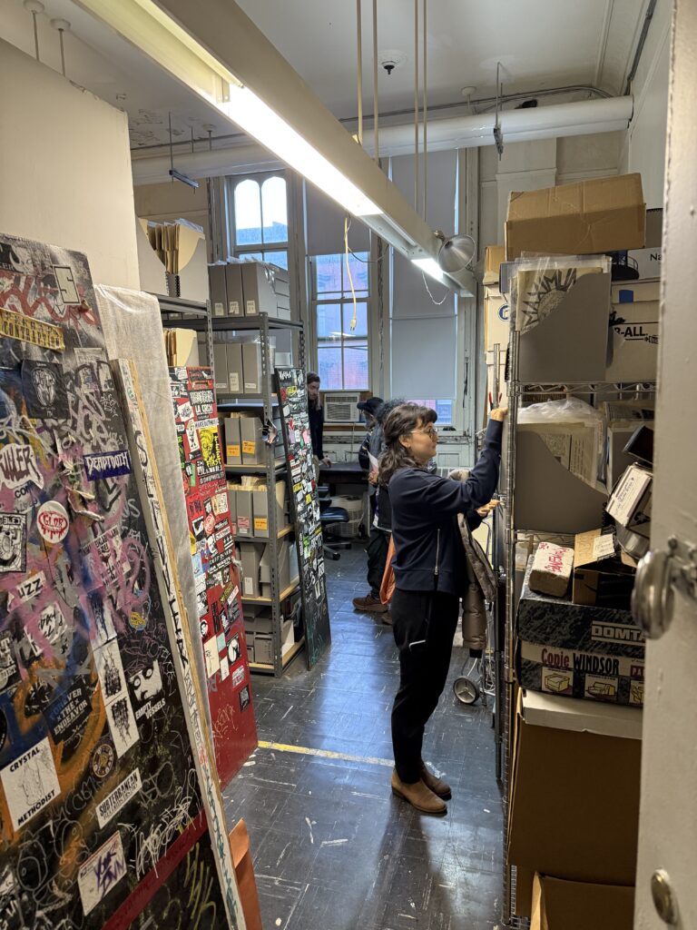 A young woman in black pants and blue jacket exploring folders full of zines on a metal rack; to the left: stacks zines and little magazines, with endcaps featuring graffiti and punk stickers 