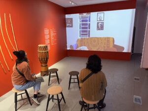 Two people sit on stools in a room with red walls, watching a screen in front of them