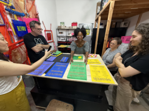 A wide-angle shot of people gathered around a work table. Zoe Pulley, a Black woman wearing checkered dress, stands in the center holding a green page from a zine.