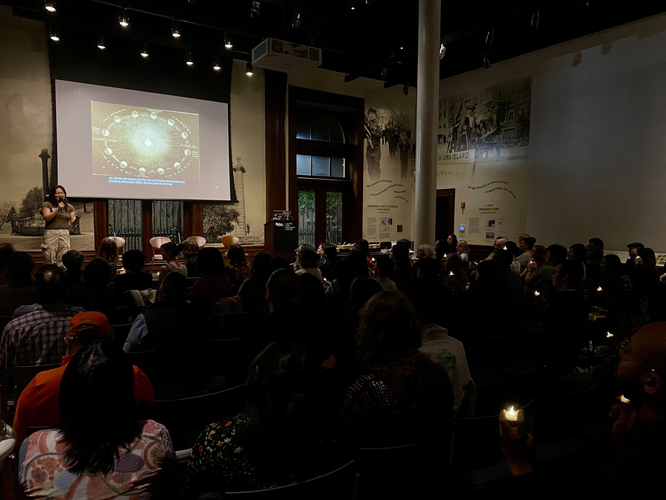 A crowd sits in a dark room with a single figure on a stage. Small lights are scattered through the crowd and an astronomy illustration is projected onto a screen behind the speaker, setting a calm, reflective mood.